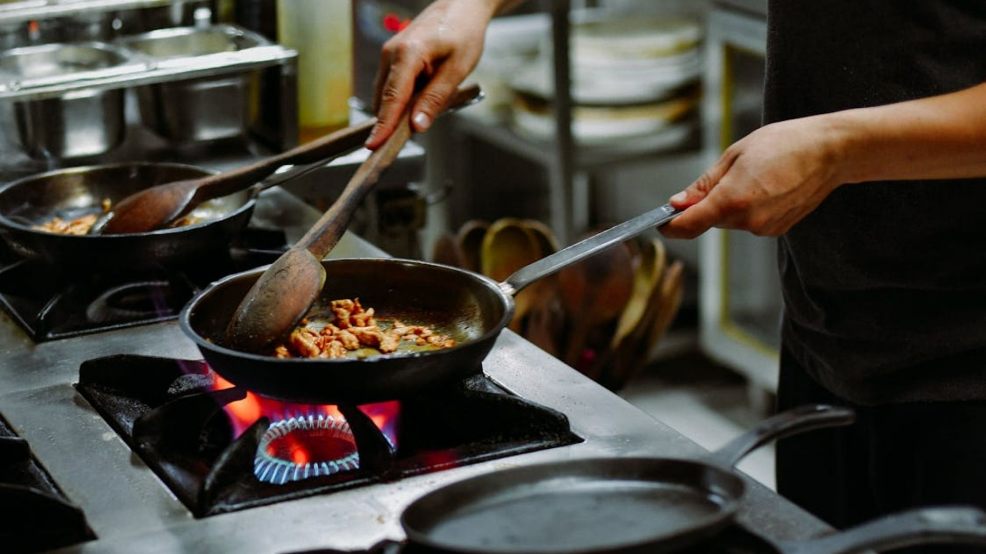 A person cooks meat in a frying pan over a gas stove, using a wooden spoon. The kitchen setting is busy, conveying a sense of focused culinary activity.