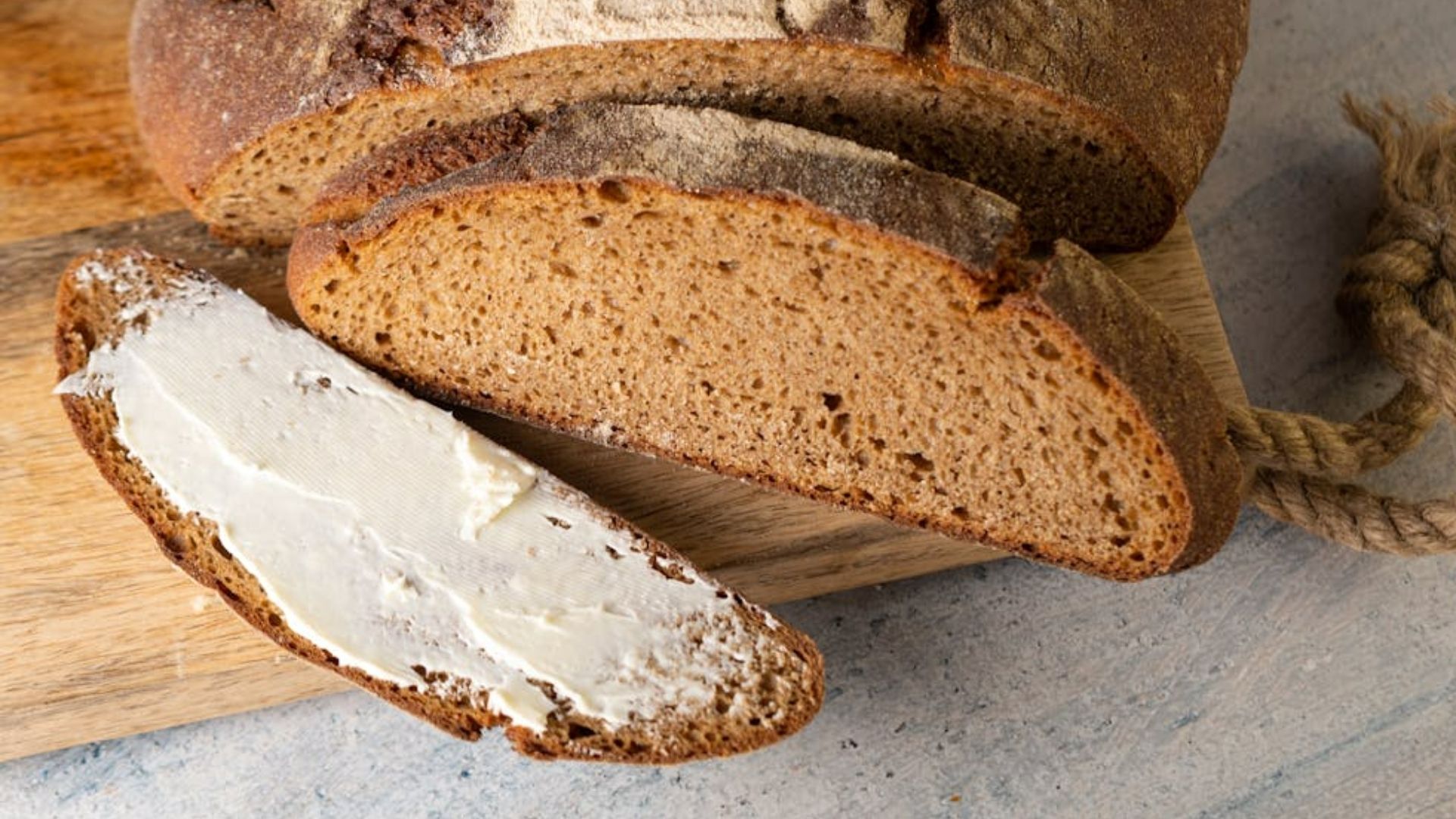 A loaf of rustic brown bread with a slice cut off, spread with creamy butter, rests on a wooden board. The setting conveys a warm, inviting feel.