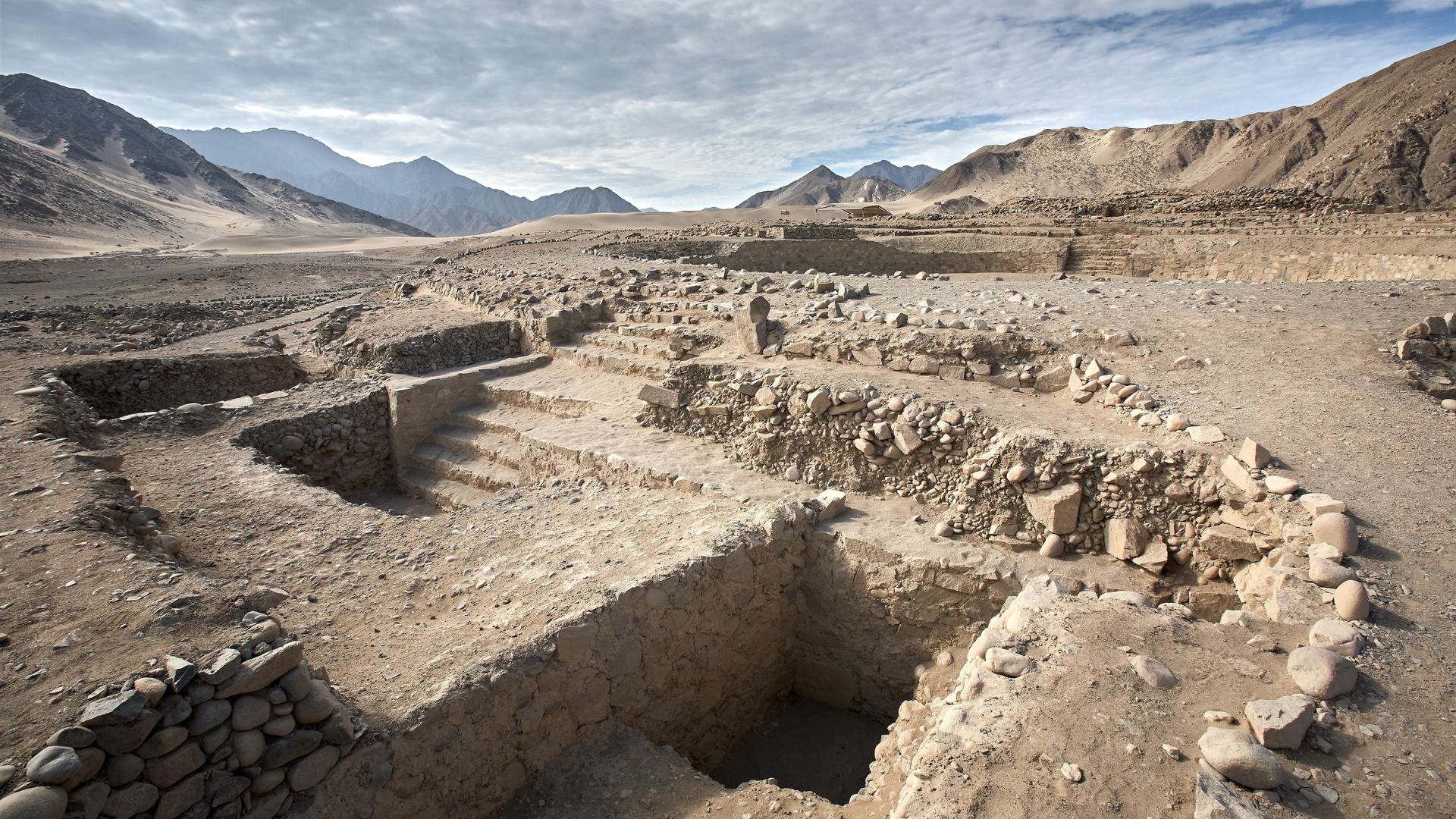 Ancient stone ruins and excavated structures in a desert landscape surrounded by mountains.