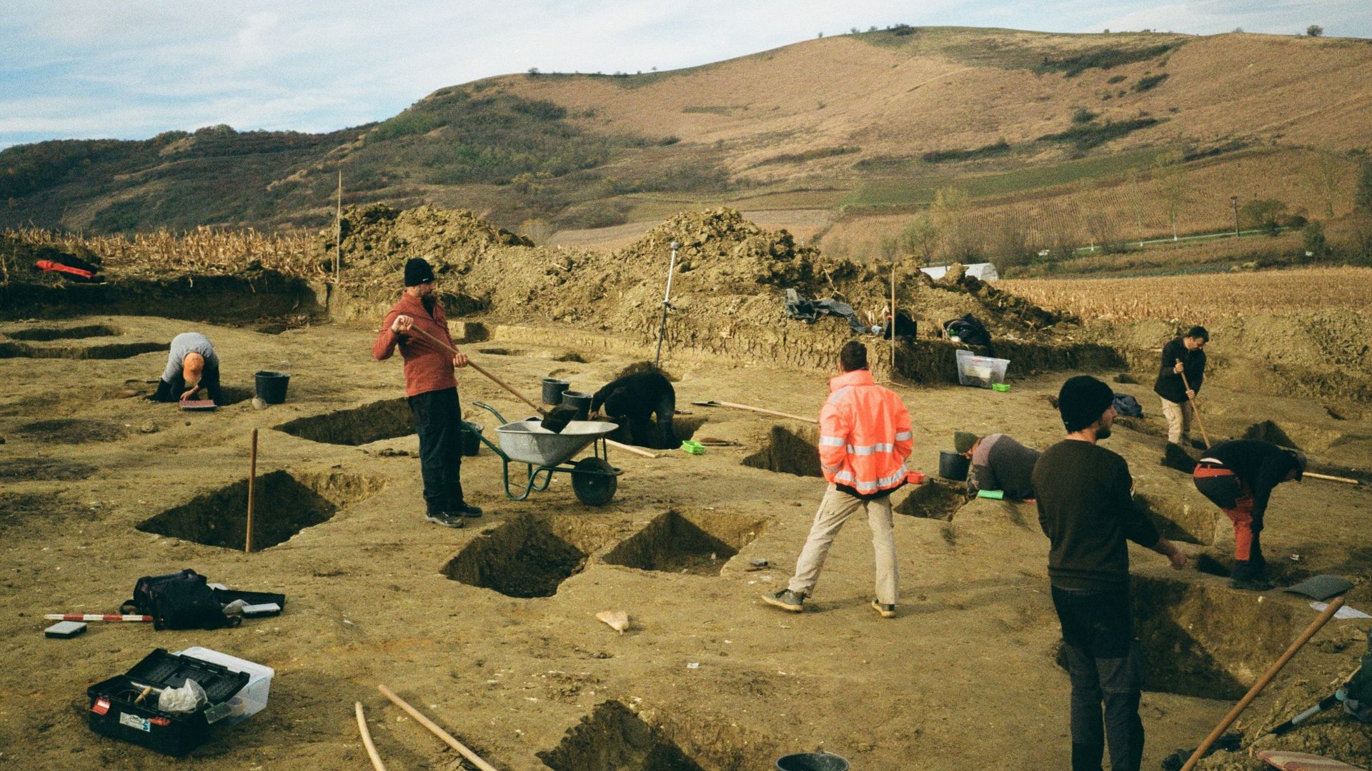 Archaeologists digging at an open field excavation with tools and trenches