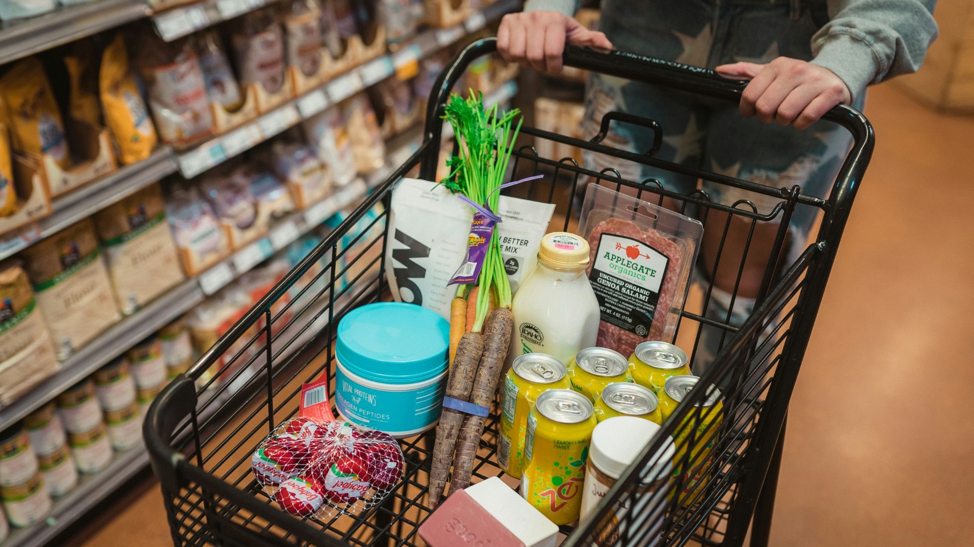 Person pushing a shopping cart full of groceries down a supermarket aisle.