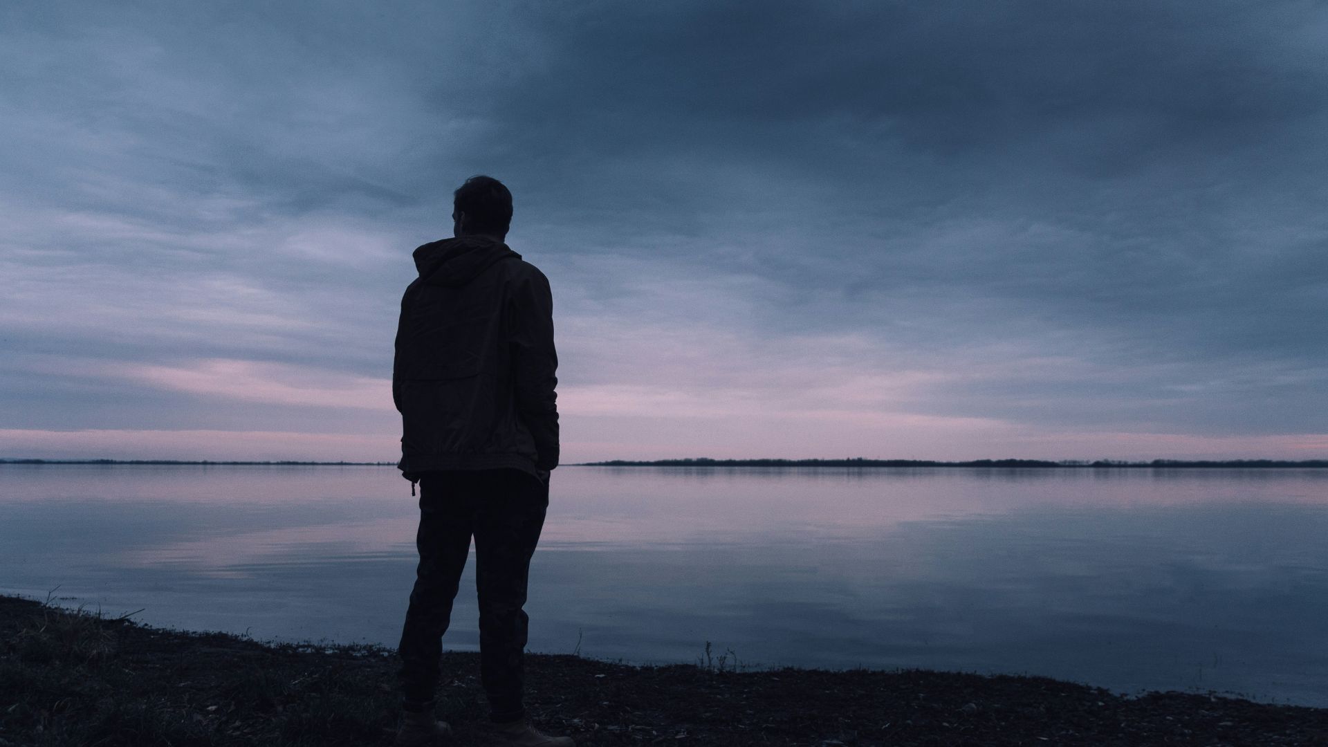 Silhouette of a man standing alone beside calm water during sunset