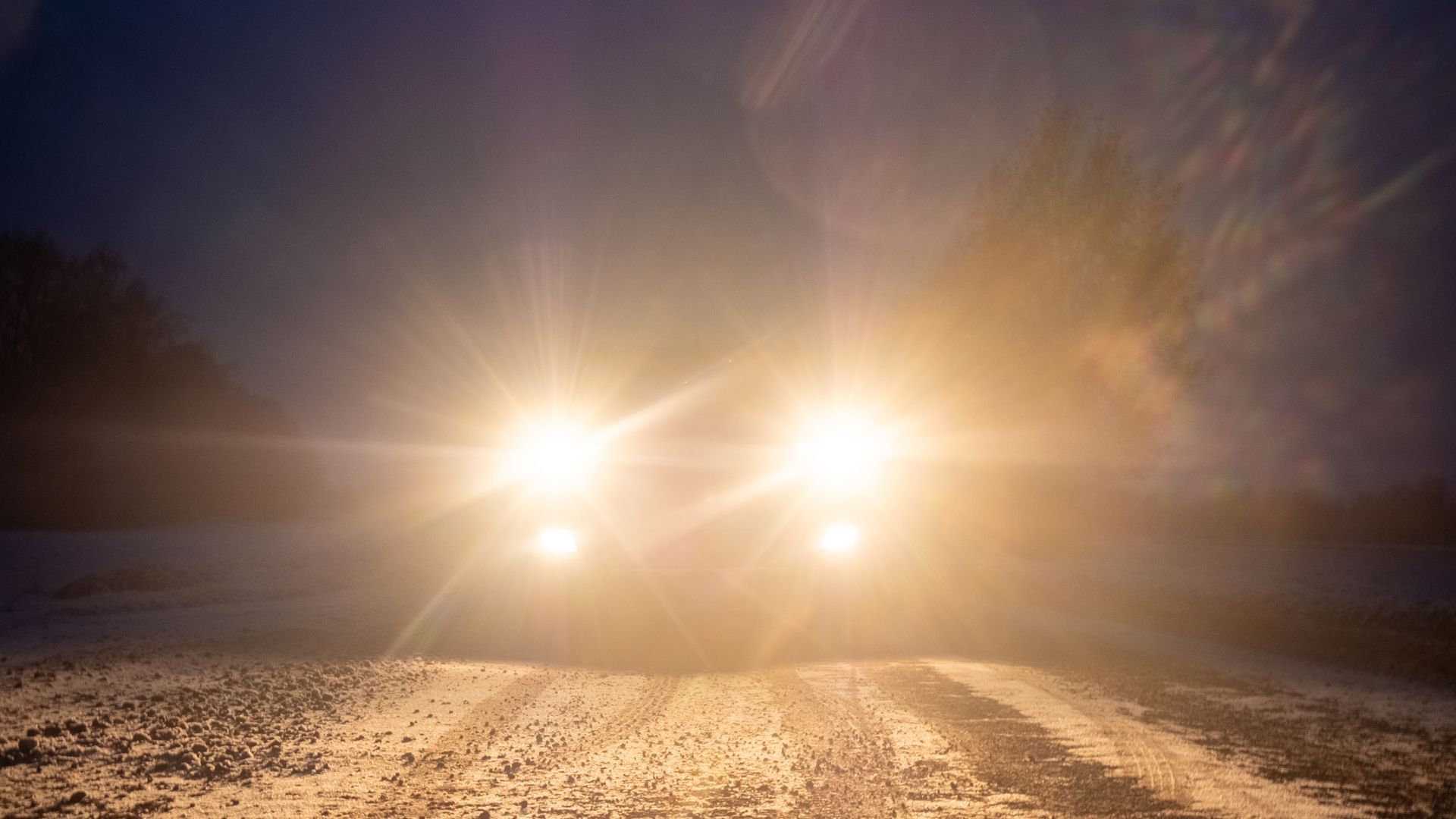 Bright car headlights shining directly at the camera on a dark, snowy road.