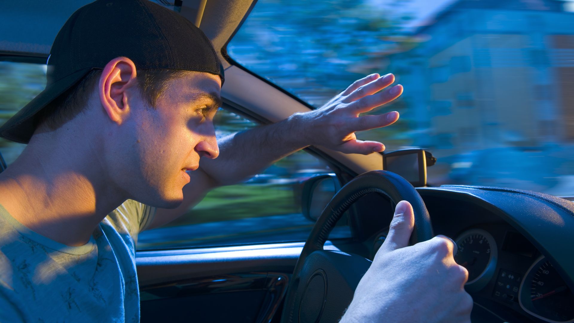 Man driving a car with one hand raised to block glare from bright light.