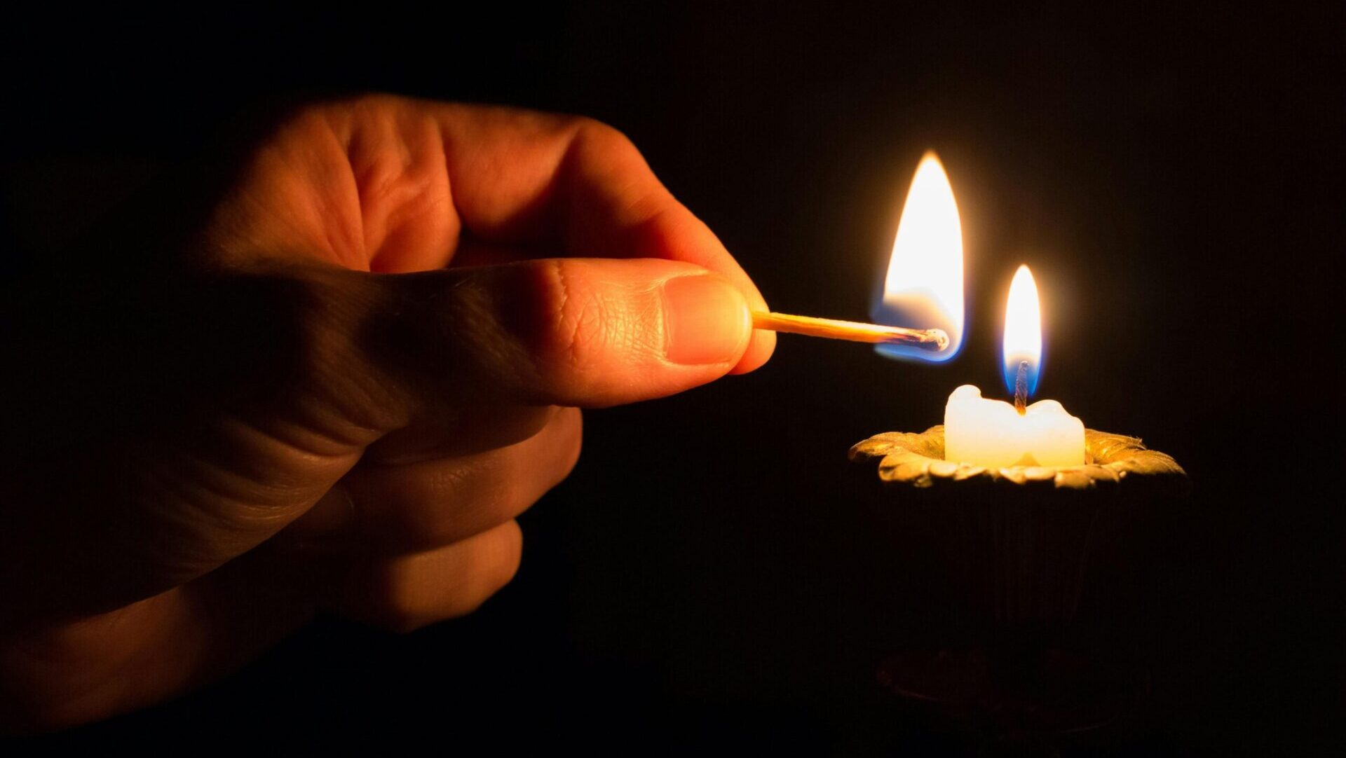 Person holding a lit matchstick in front of a burning candle, capturing the flame and warm glow.