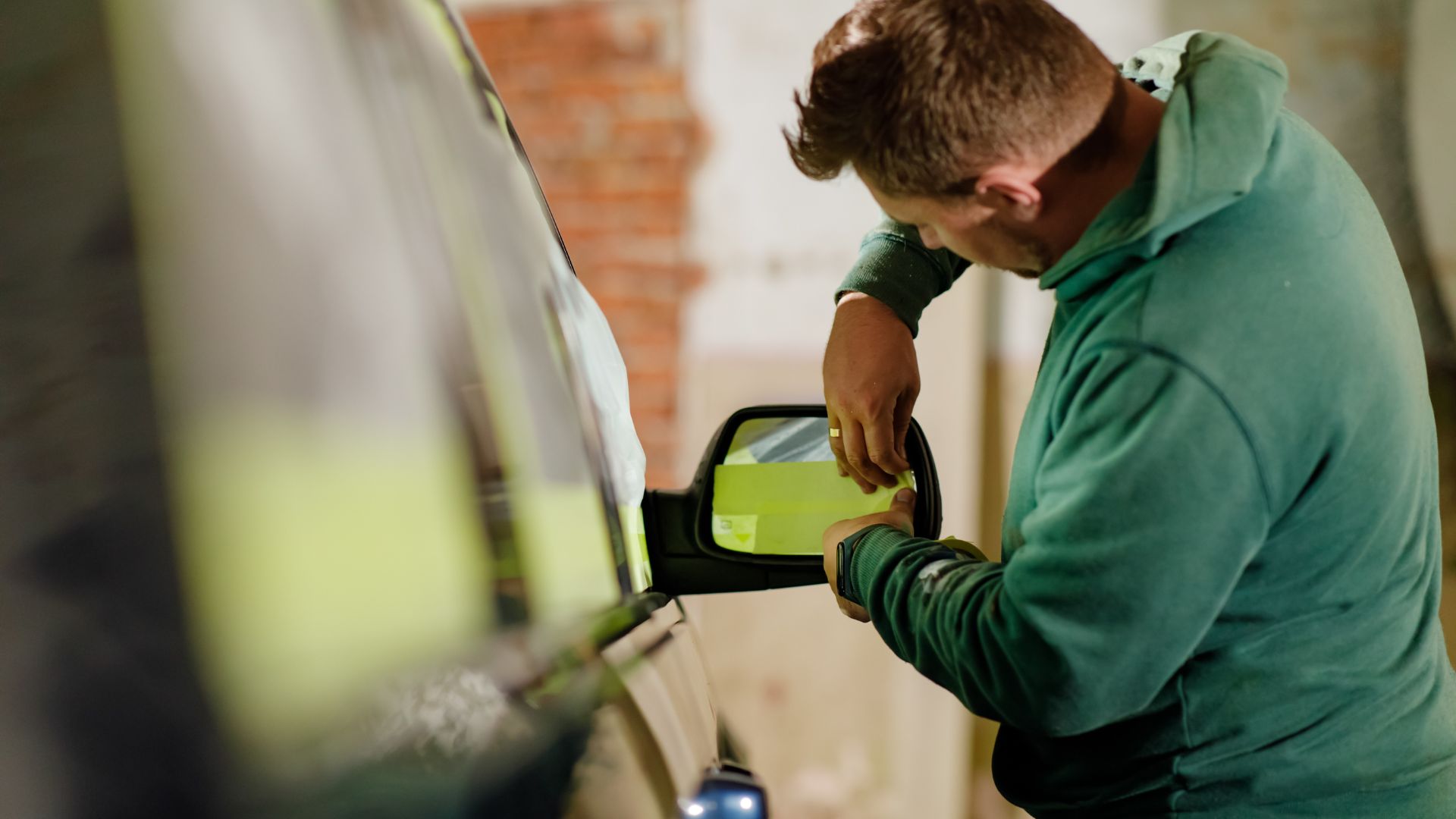 Man applying yellow tint or film to a car’s side mirror in a garage.