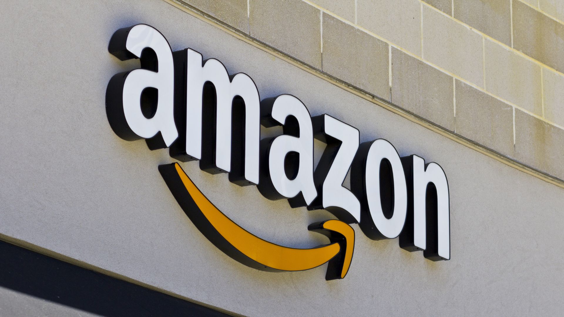 Amazon logo mounted on the exterior wall of a building. The black and white lowercase lettering includes the curved yellow arrow beneath the wordmark that points from the letter “a” to “z.”