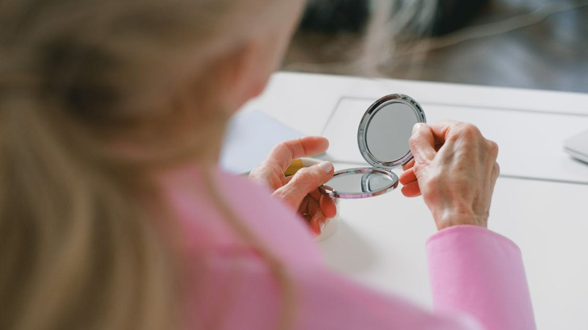 An elderly person in a pink shirt holds an open compact mirror