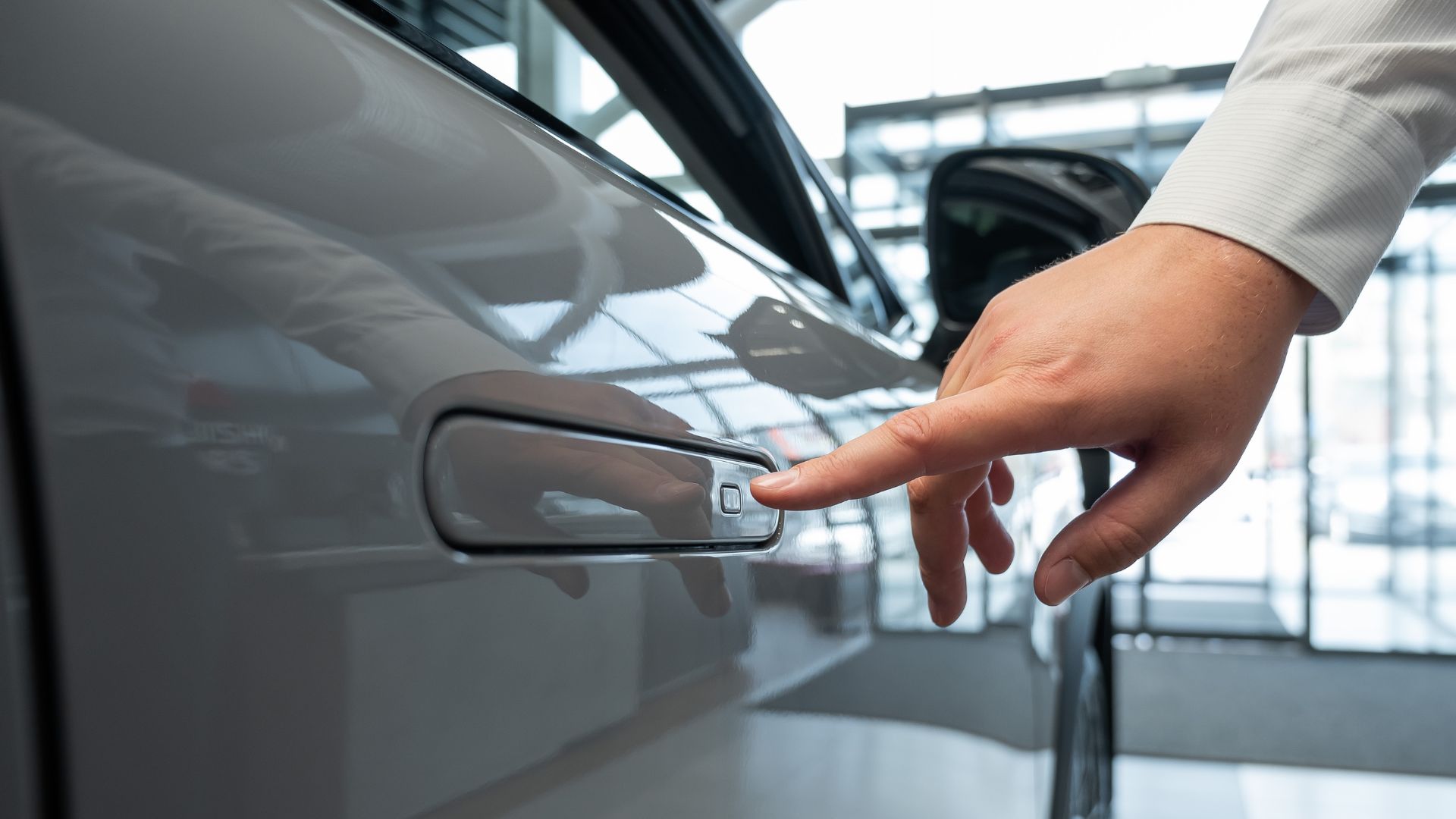 A close up of a hand pressing a flush car door handle on a modern gray vehicle inside a showroom.