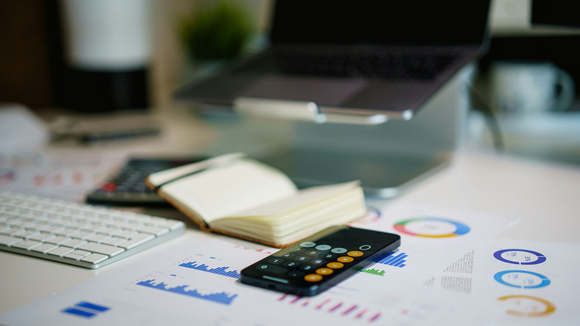 Calculator and smartphone resting on financial charts beside a laptop and notebook.