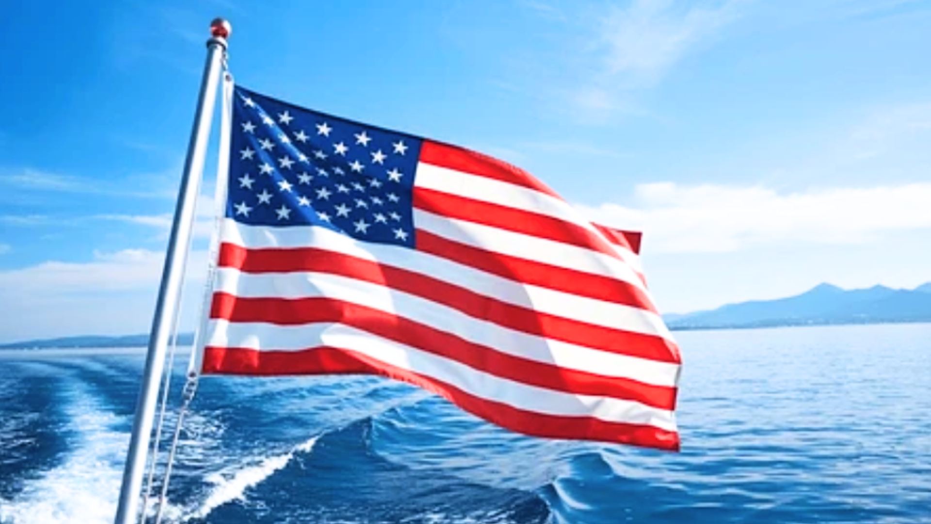 American flag flying on a boat with ocean and mountains in the background.