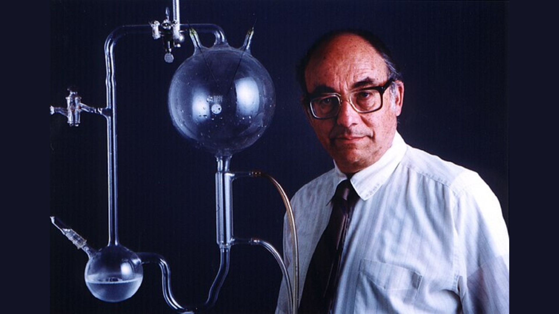 Stanley Miller stands beside a large, clear glass laboratory flask apparatus with multiple round bulbs and connecting tubes mounted on a metal stand.