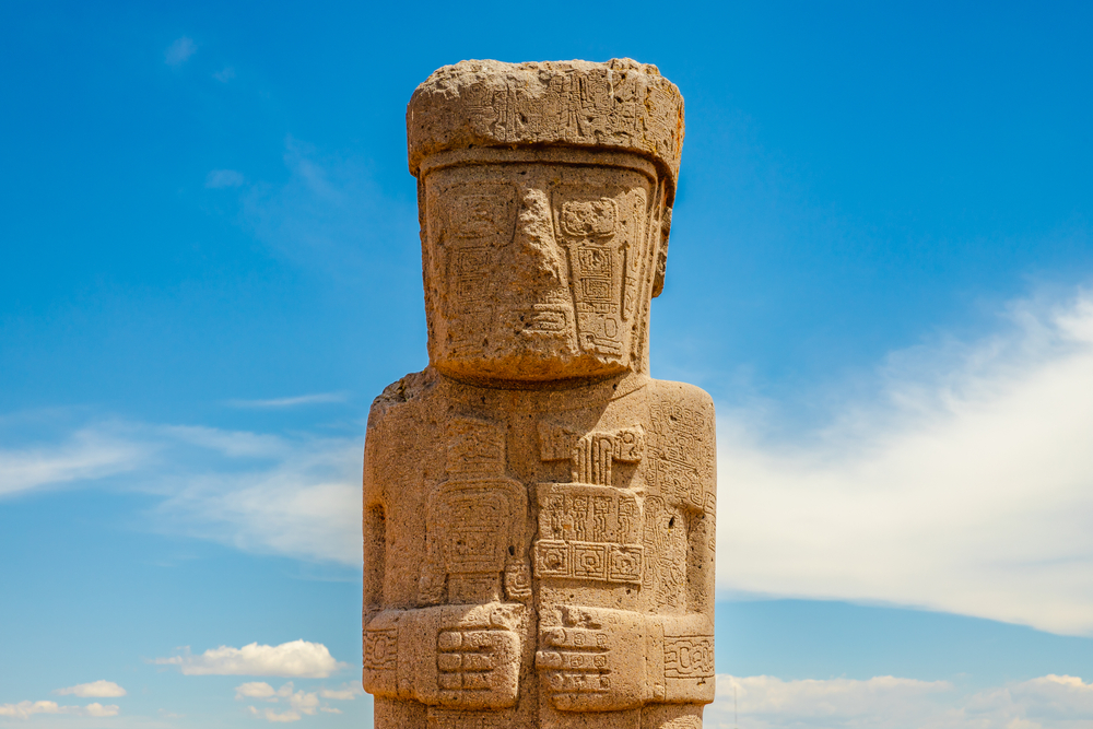A carved stone monolith from Tiwanaku standing against a blue sky, featuring intricate geometric and symbolic designs.