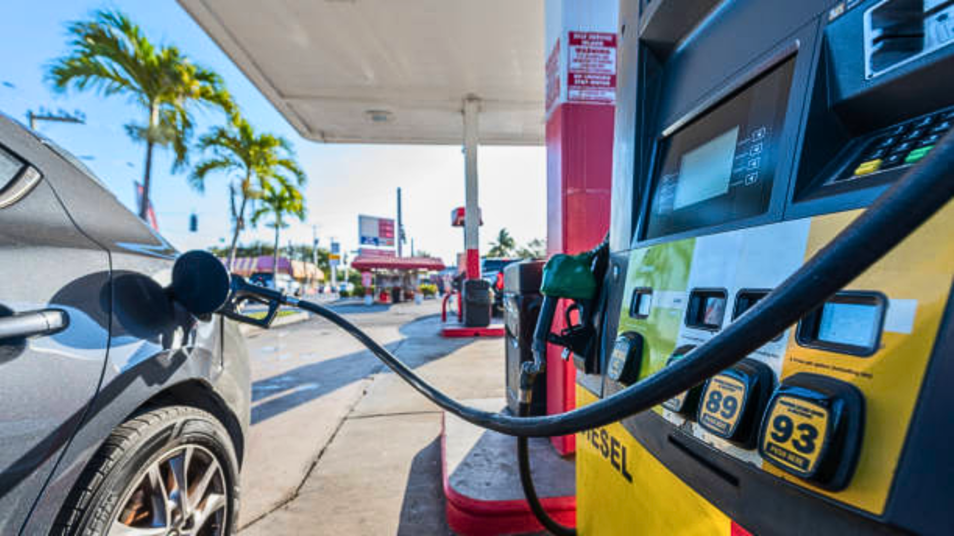 Close-up of a fuel nozzle inserted into a car at a gas station, with gas pumps and palm trees in the background.