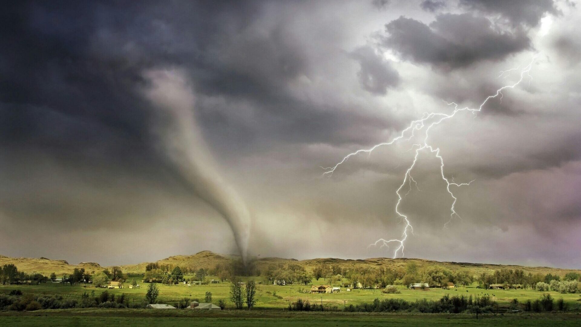 Lightning striking near a tornado funnel approaching a small village.