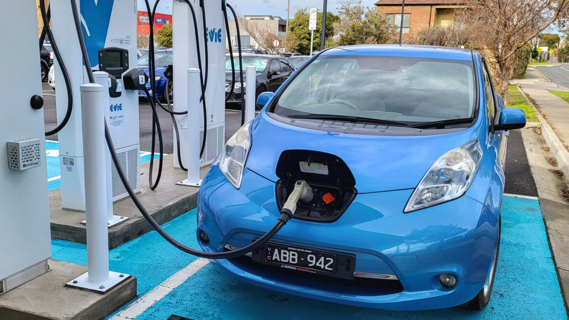 A blue Nissan Leaf electric car parked at a public charging station with the charging cable connected to the front port of the vehicle.