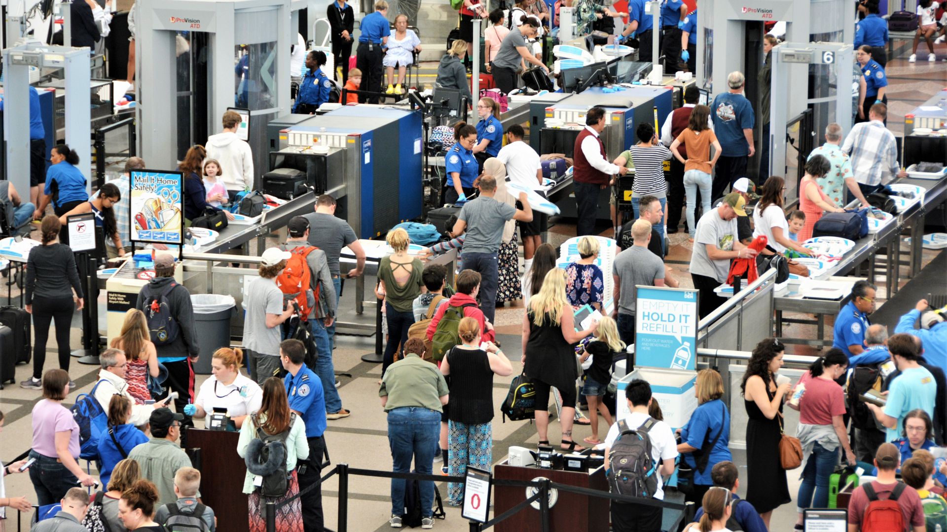 Crowded airport security checkpoint with TSA agents screening passengers and luggage at multiple lanes.