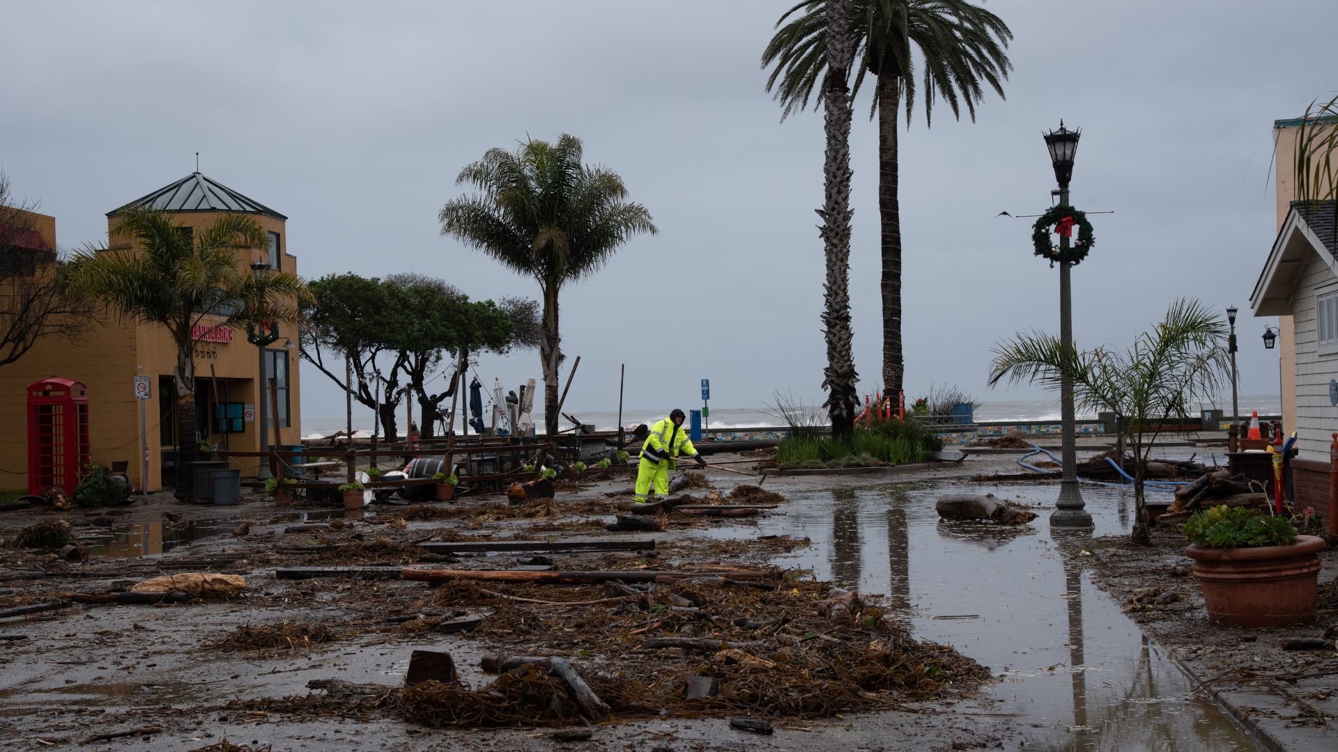 Worker clearing storm debris from flooded coastal street lined with palm trees in Santa Cruz.
