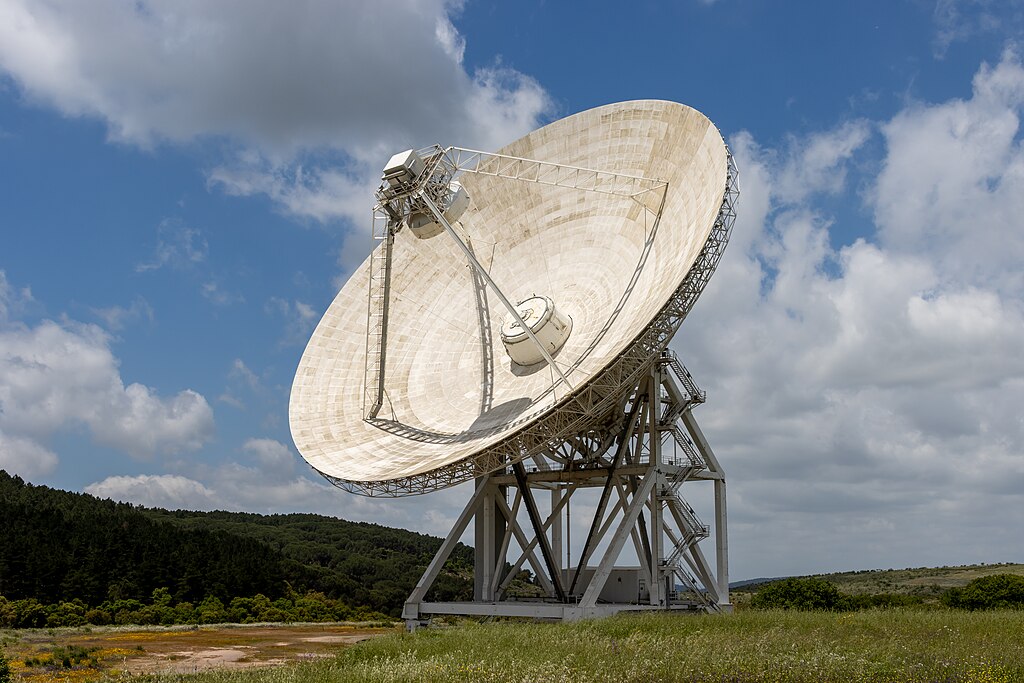 Large radio telescope dish at the Sardinia Radio Telescope observatory pointed toward the sky above a grassy landscape.