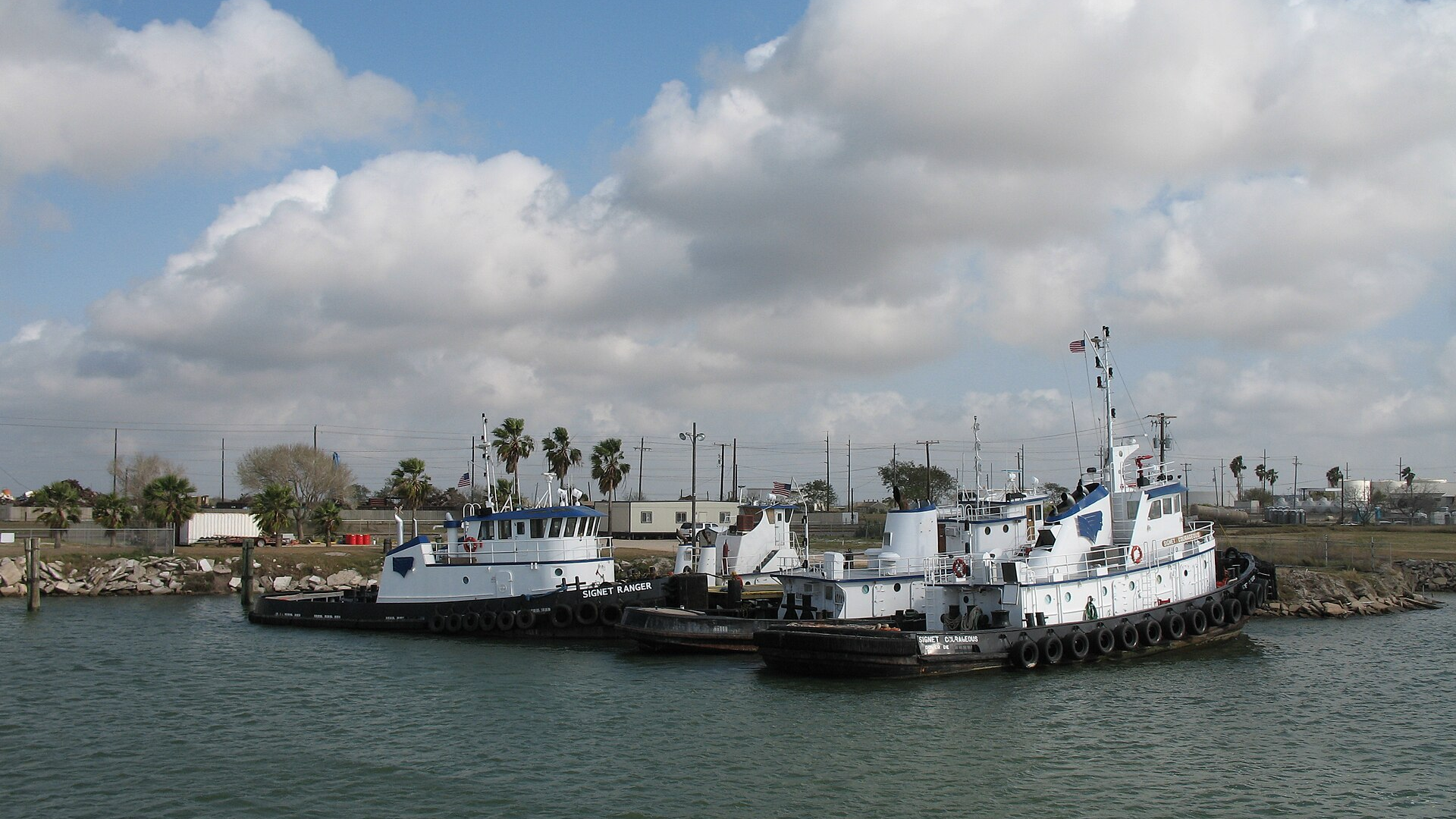 Three white and blue tugboats docked at a harbor under a cloudy sky.