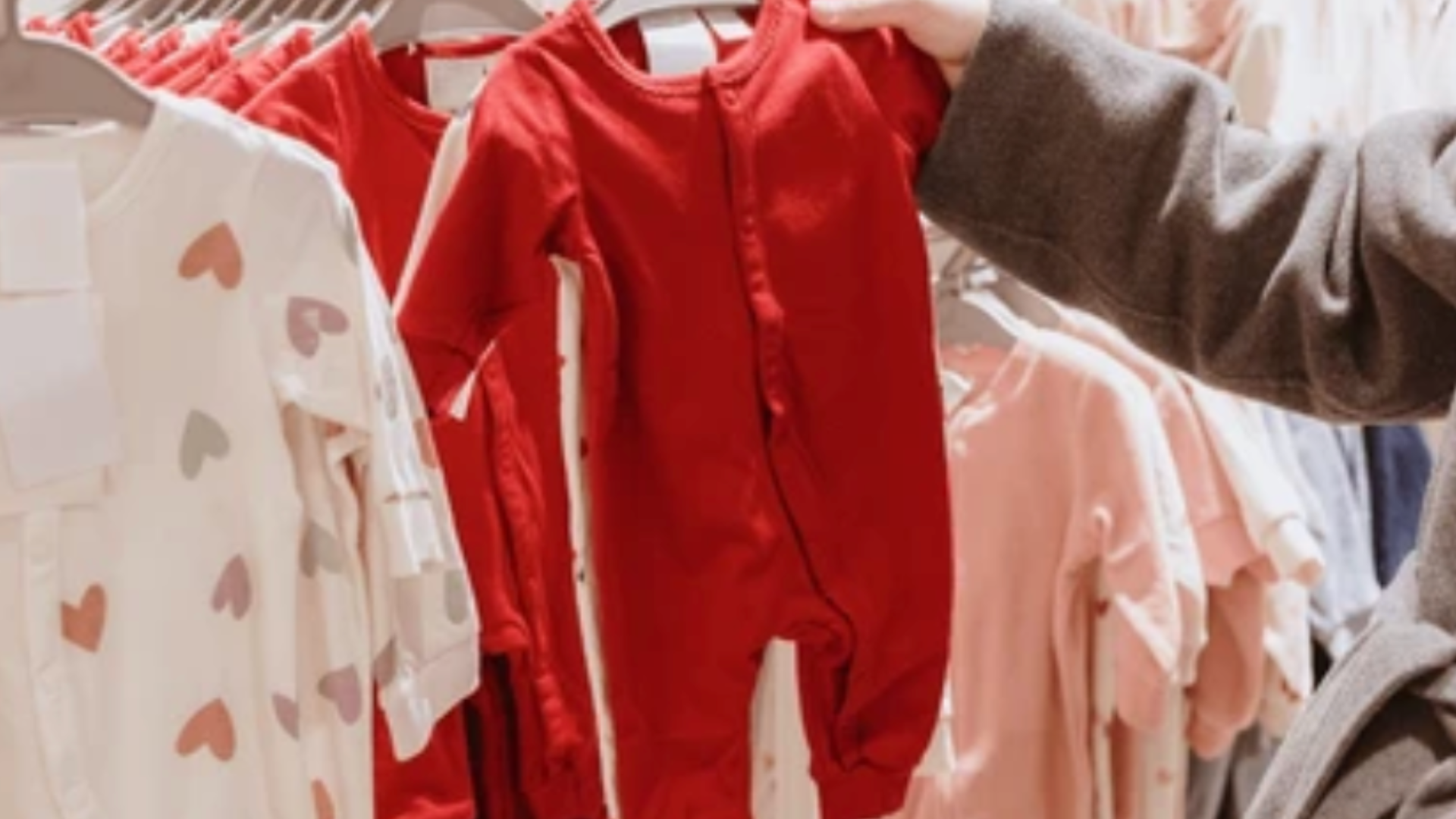 A person’s hand reaching for a bright red baby onesie hanging on a clothing rack in a retail store.