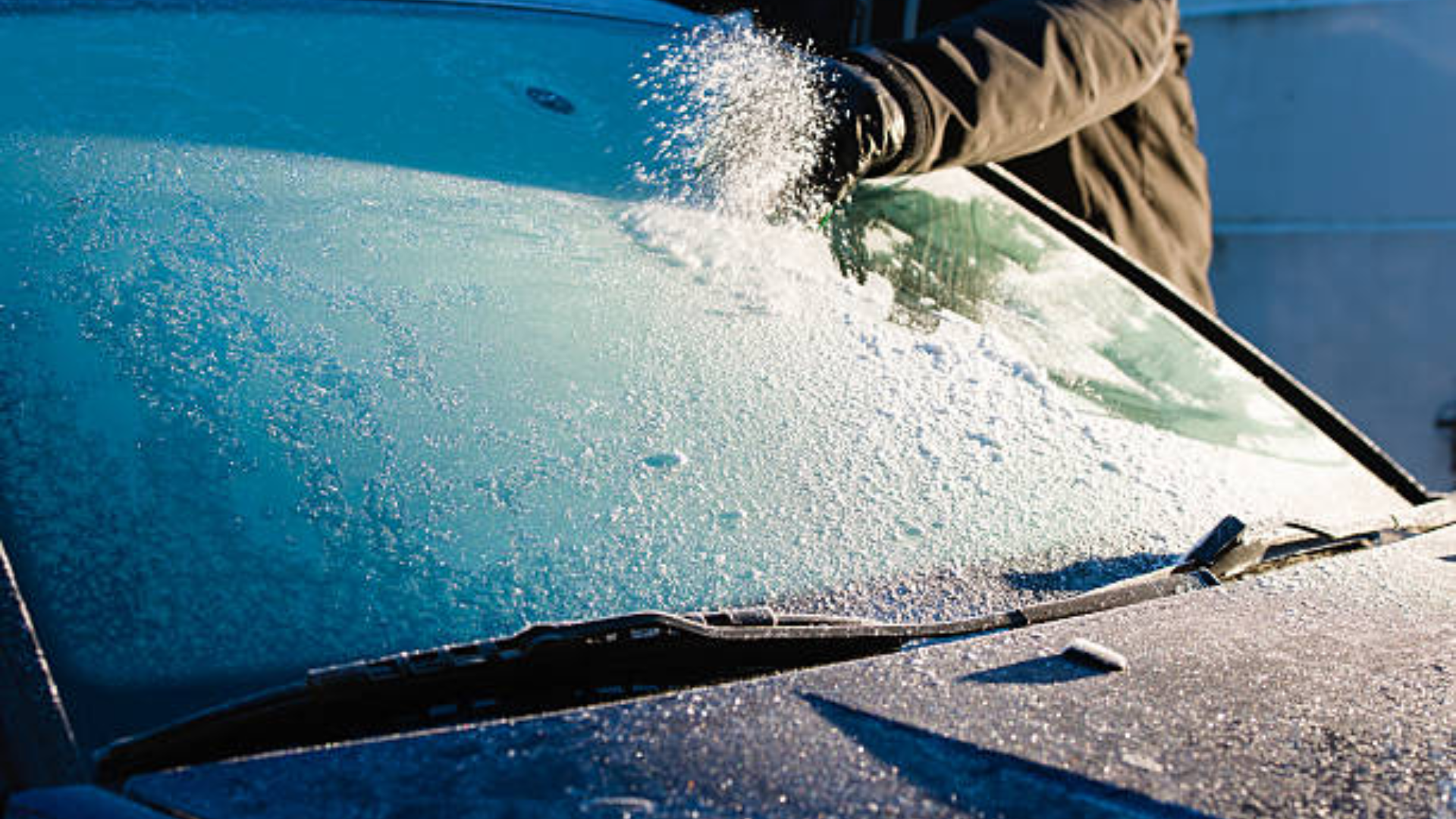 A person using an ice scraper to clear a thick layer of frost and frozen snow from a car's windshield on a cold morning.
