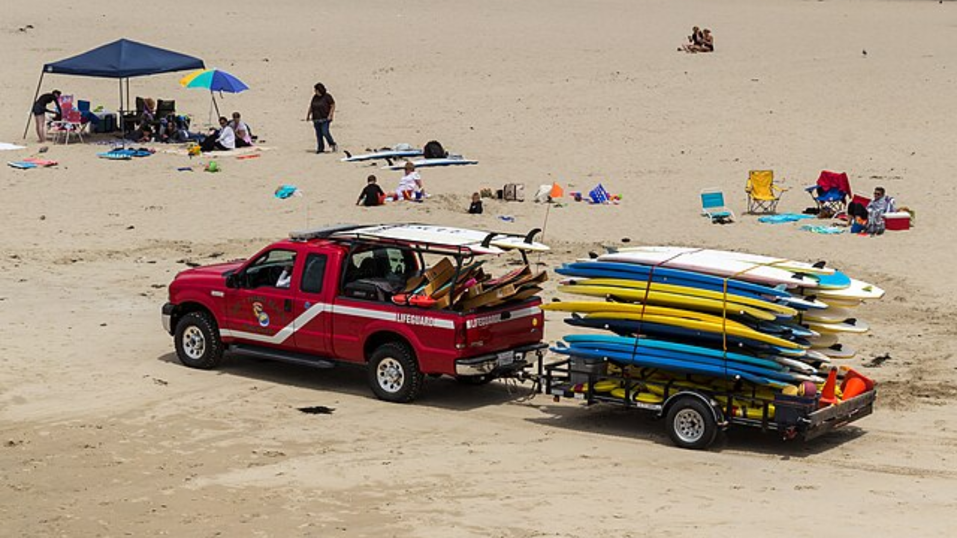 Lifeguard truck towing trailer of surfboards parked on sandy beach with people nearby.