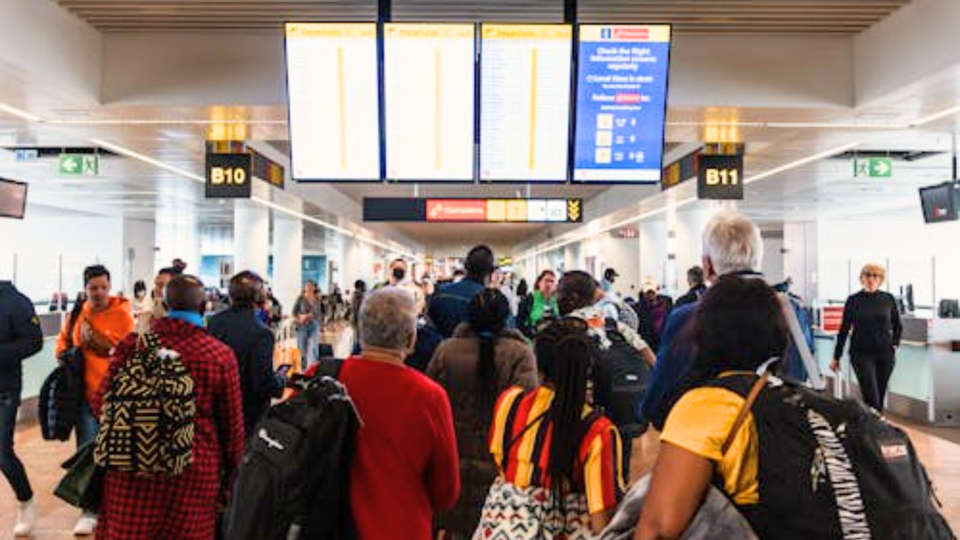 A crowd of travelers stand together looking up at bright flight information screens.