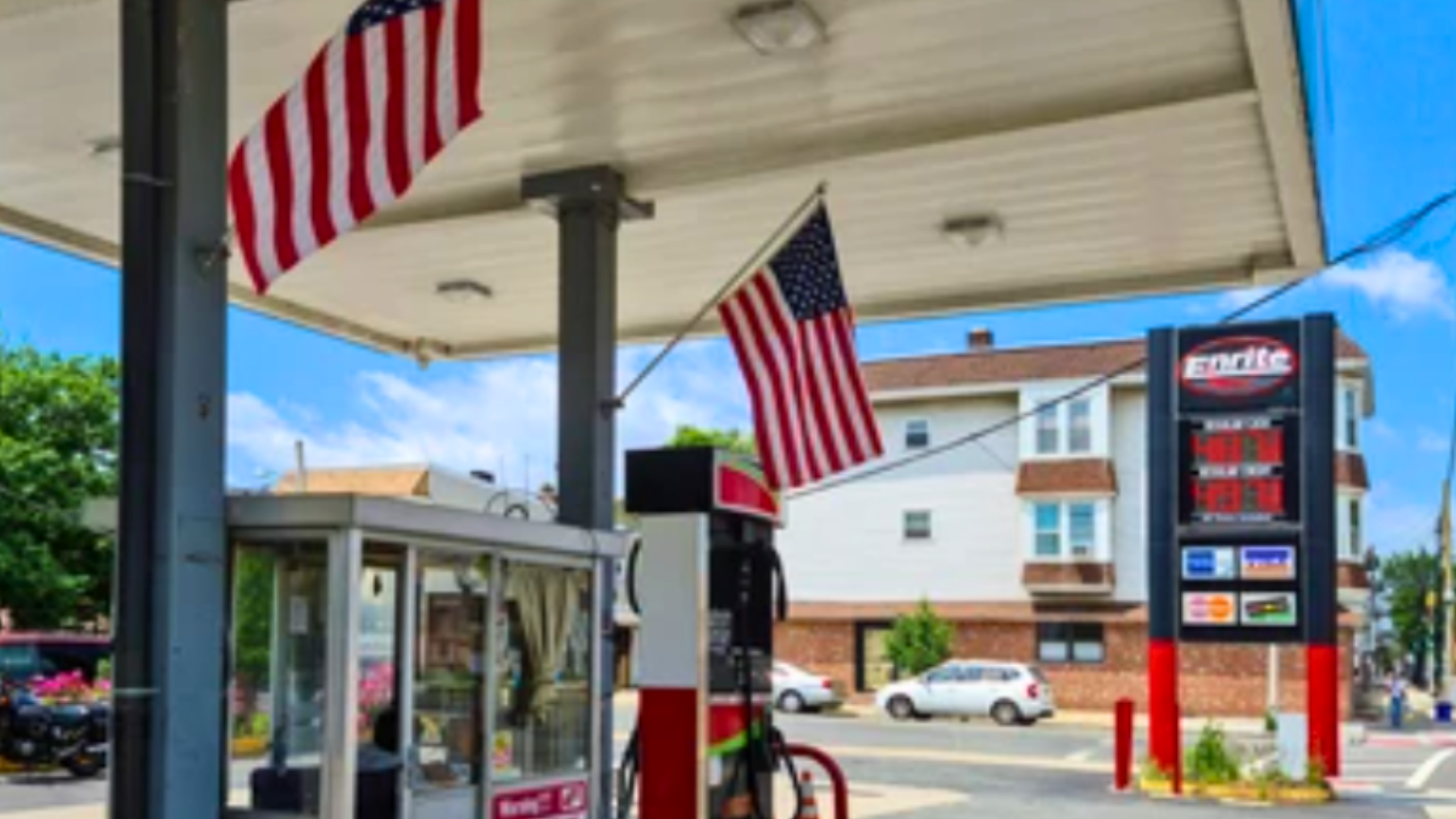 An Enrite gas station canopy decorated with American flags, showing fuel prices near a residential building.