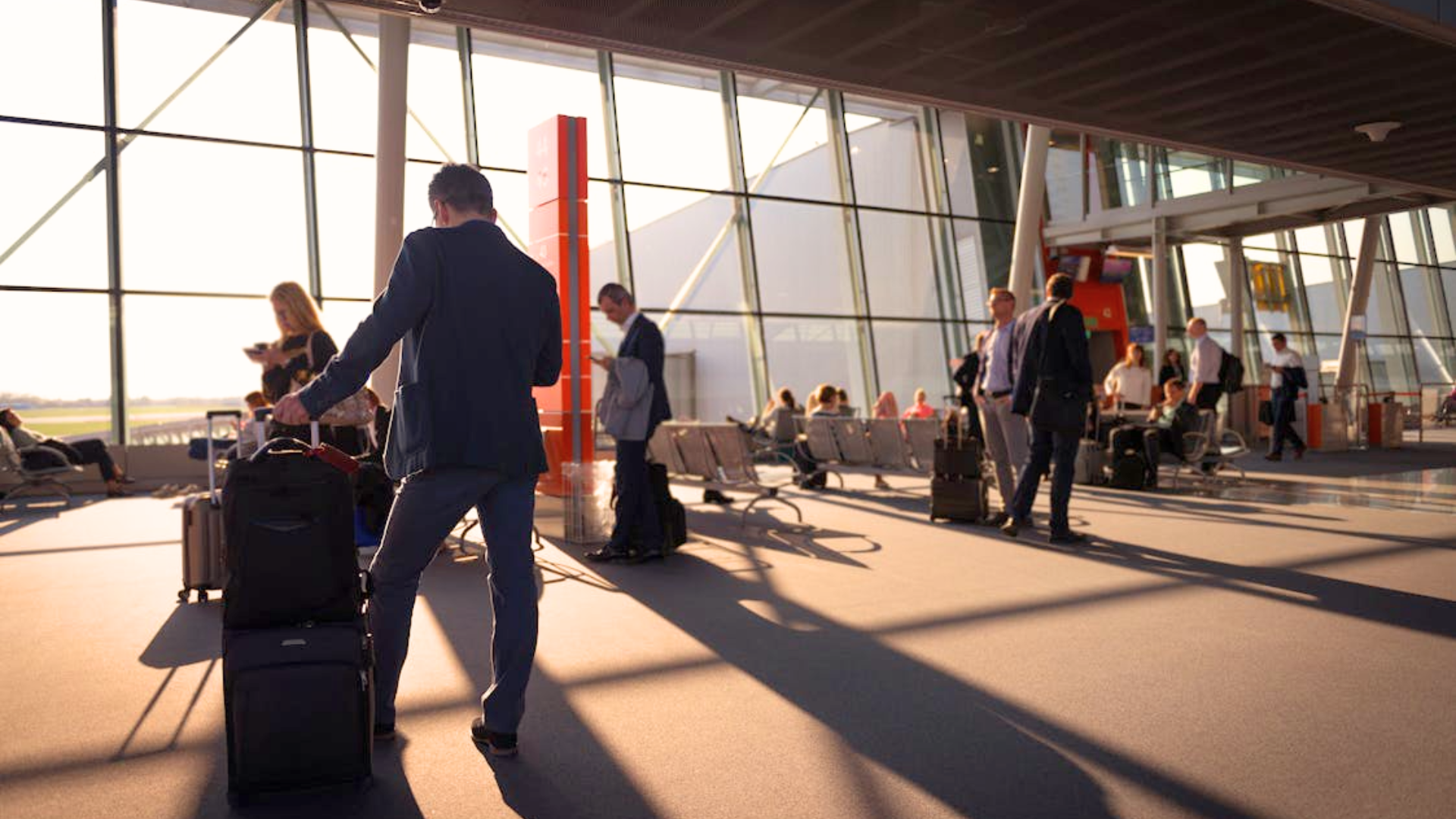 A man stands with his suitcase in a sunny airport room with large windows.