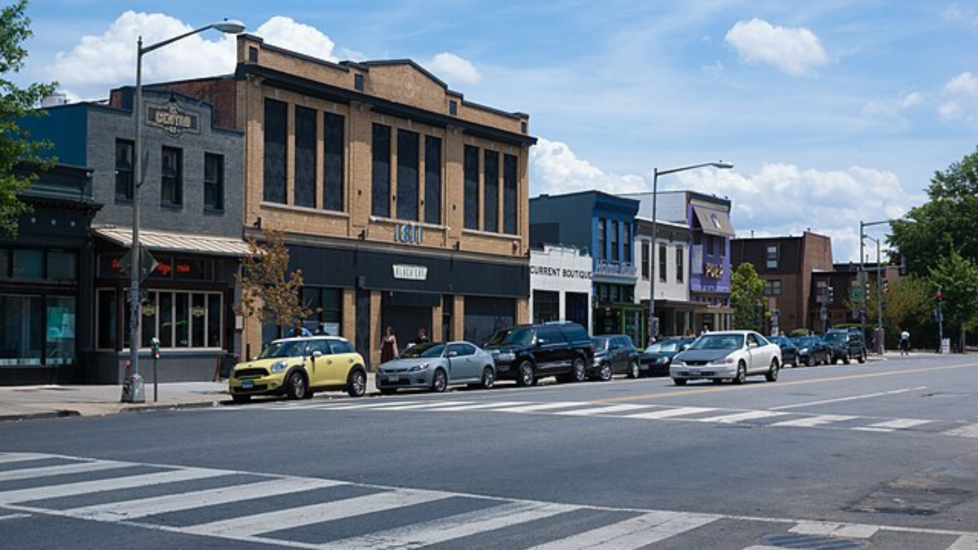 A row of colorful commercial buildings and storefronts along a city street with cars parked in front.