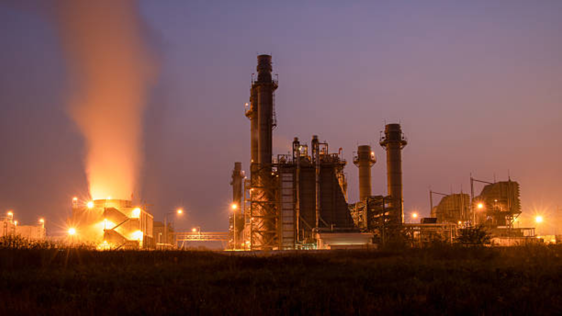 Dusk view of an illuminated industrial power plant or refinery with steam rising from a cooling tower.