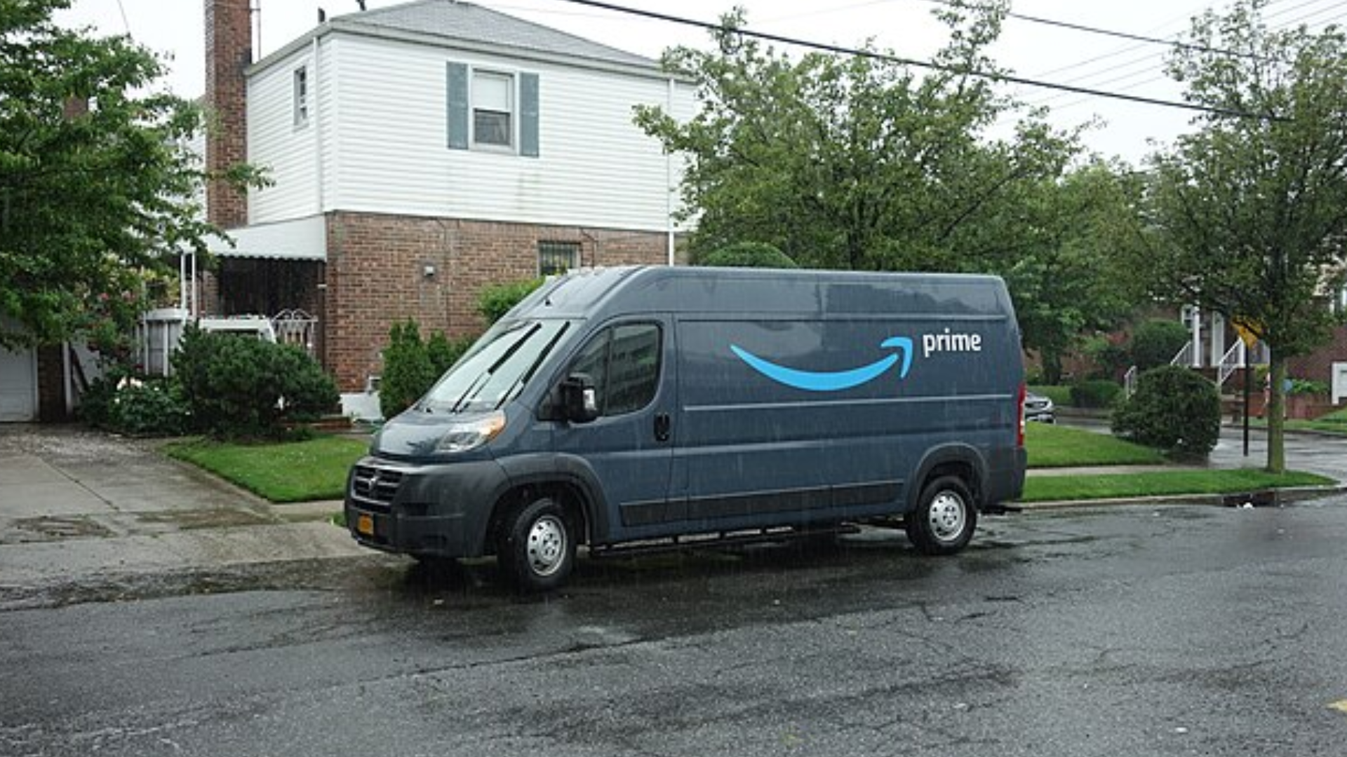 A blue Amazon Prime delivery van parked on a residential street during a rainy day.