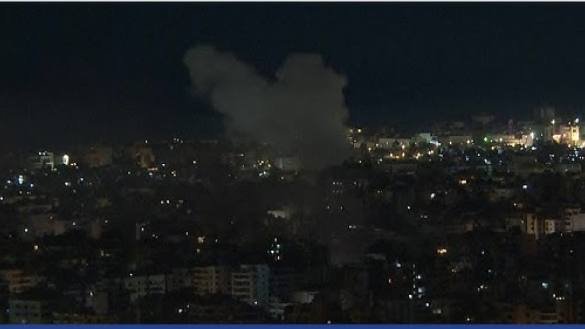 A nighttime view of a city with a large plume of dark smoke rising from a specific location amidst the city lights.