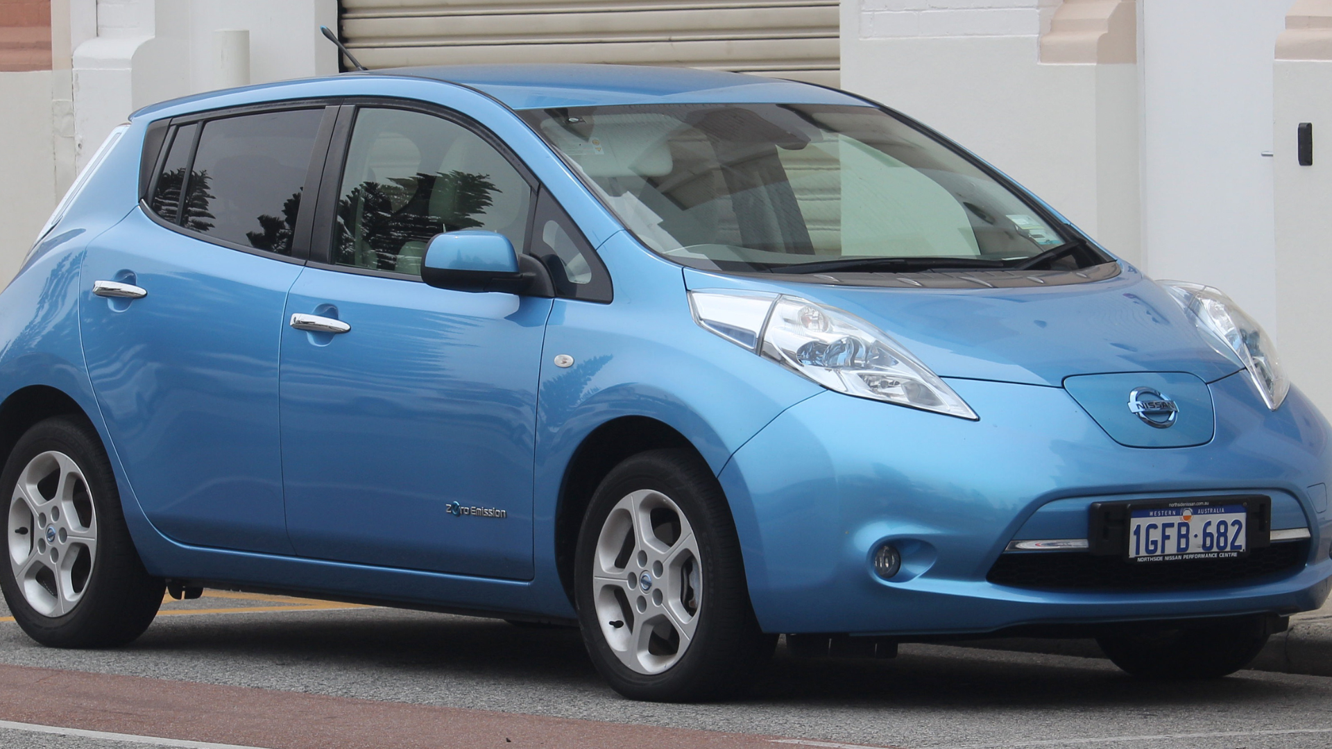 A side profile view of a light blue Nissan Leaf hatchback parked on a city street, showing the "Zero Emission" badge on the door.