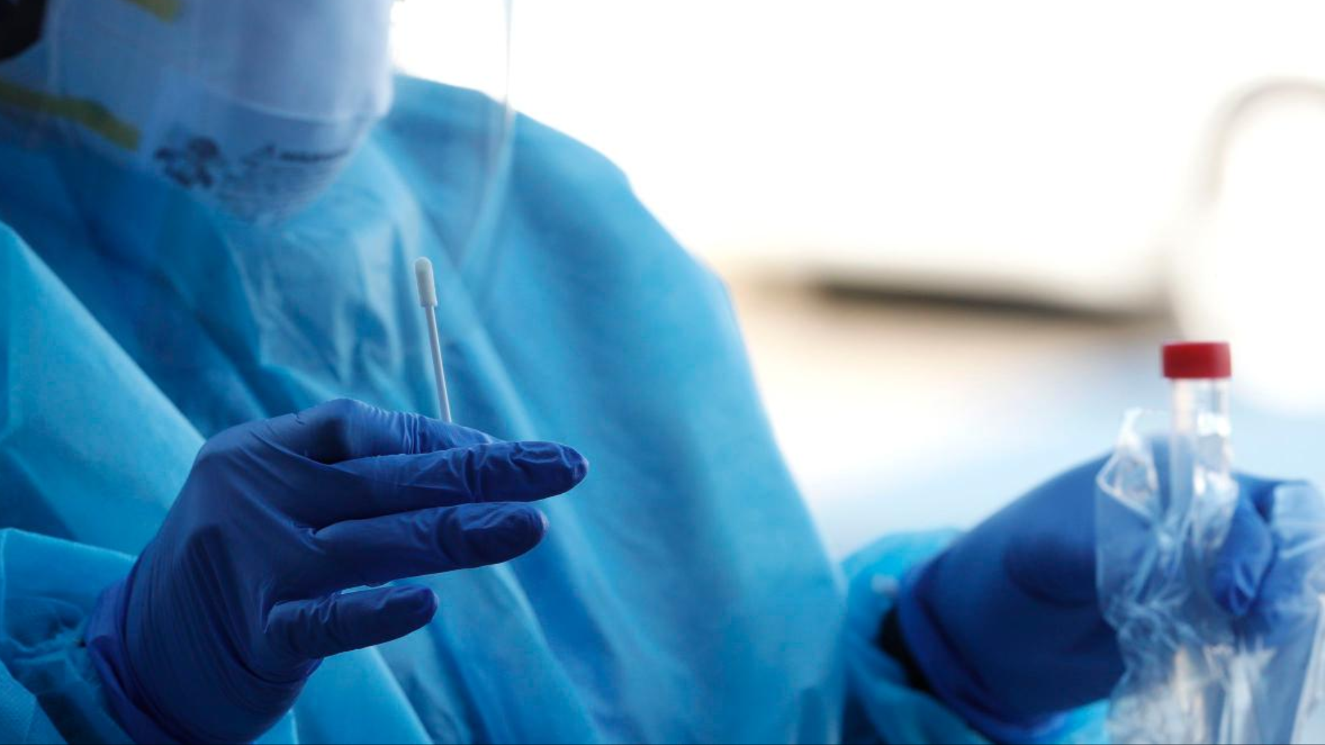 Healthcare worker holding a swab and test vial in PPE.
