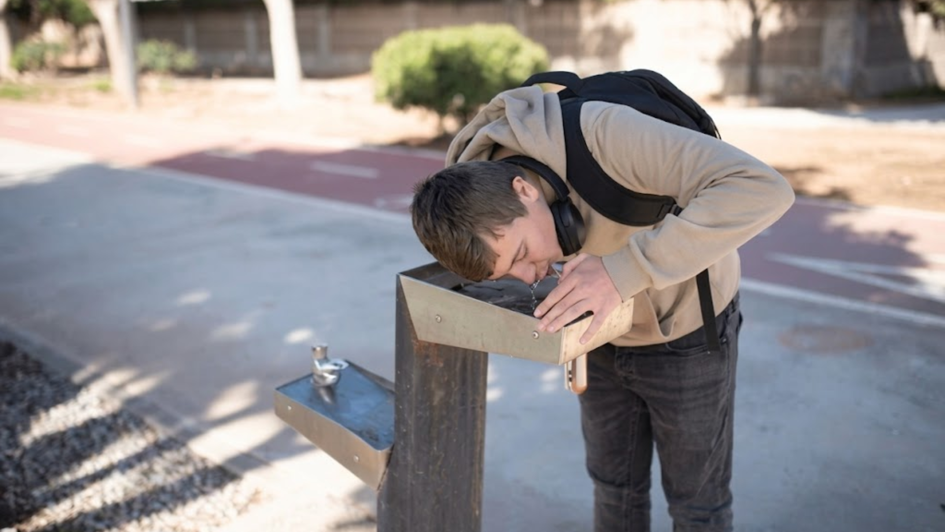 Teen drinking water from public fountain on a sunny day.