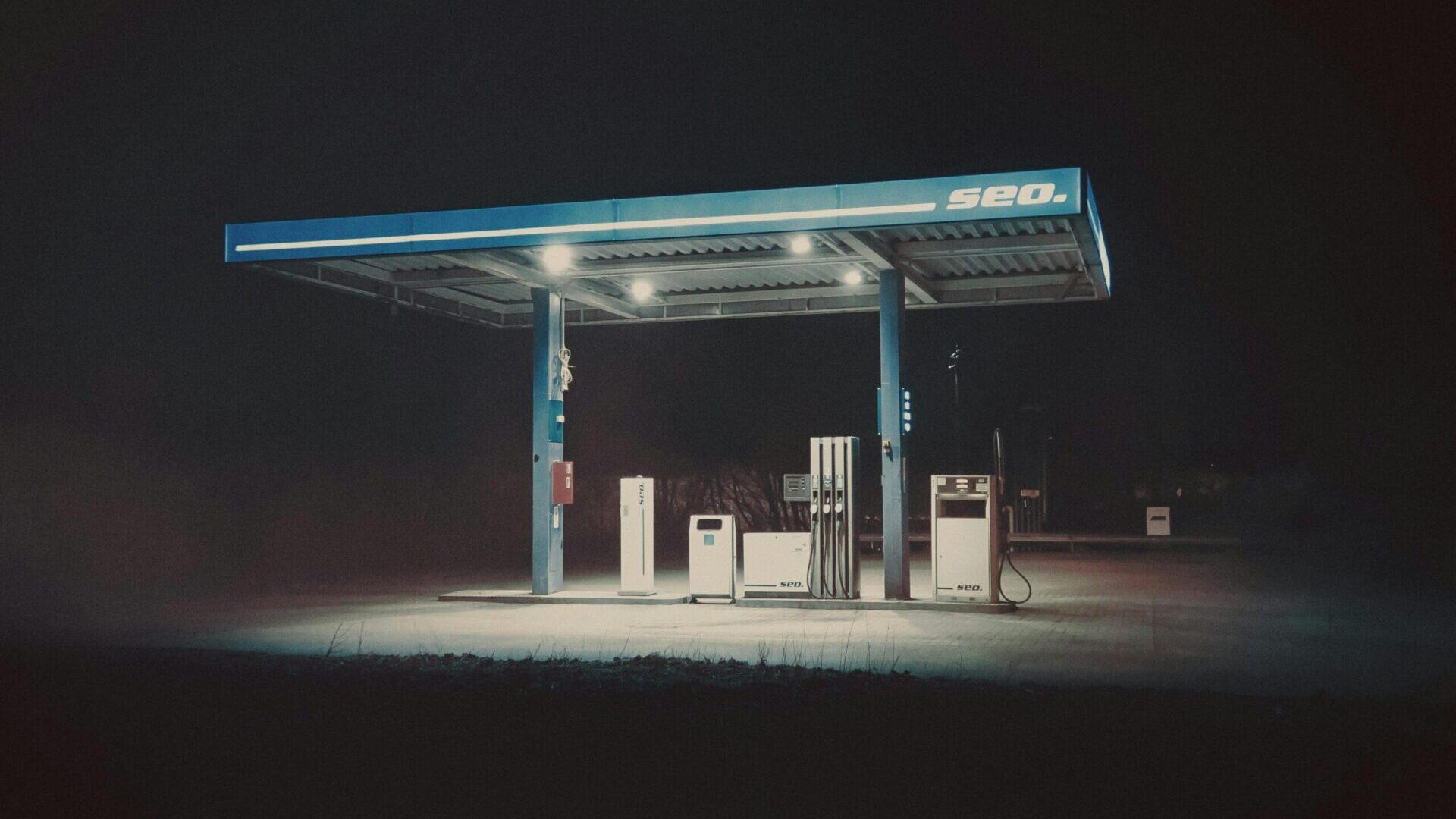 Gas station at night with illuminated pumps under a canopy in a dark setting.