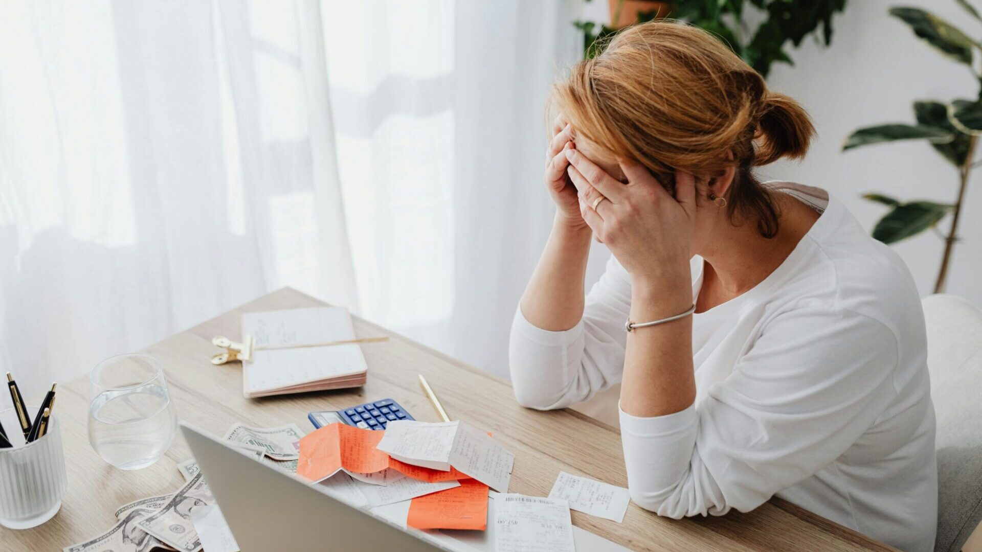 Woman sitting at a desk holding her head while looking stressed over bills and paperwork.