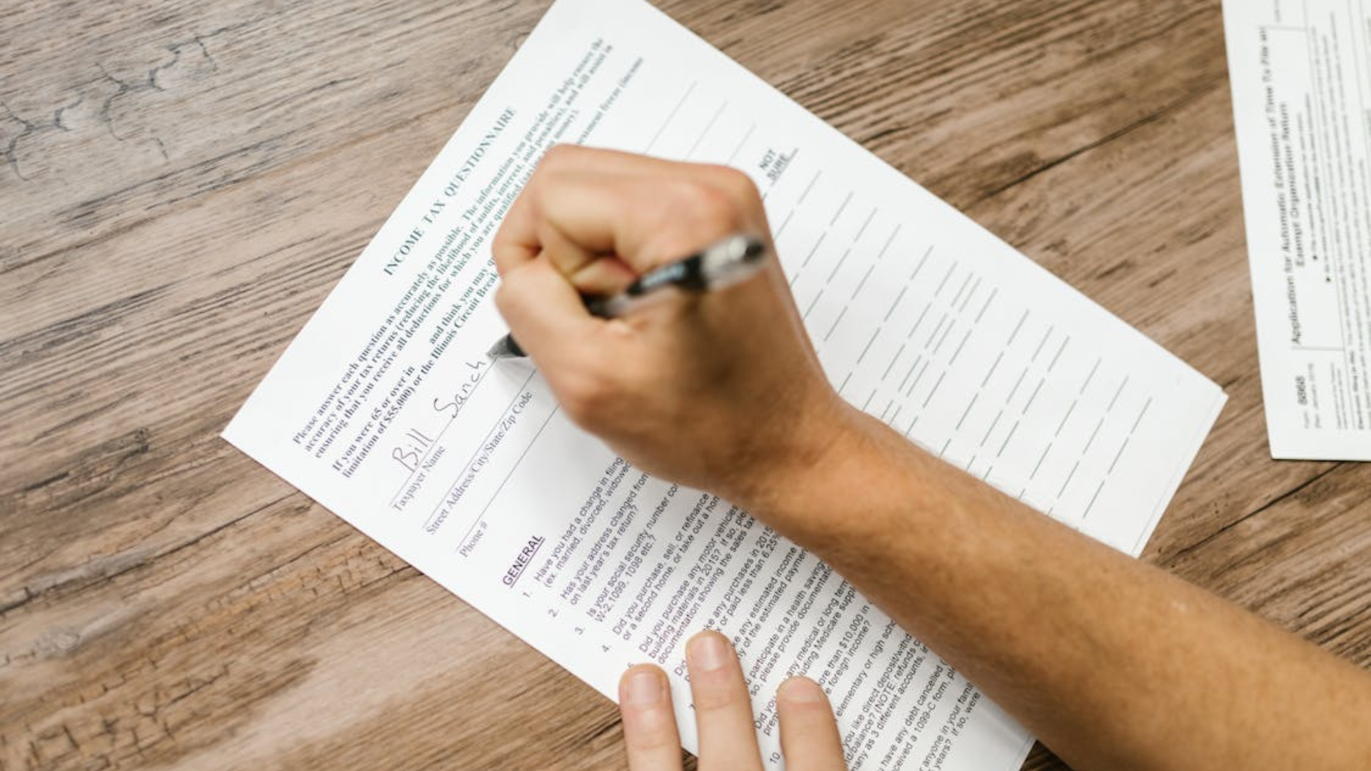 A person uses a pen to fill out an income tax paper on a wooden desk.