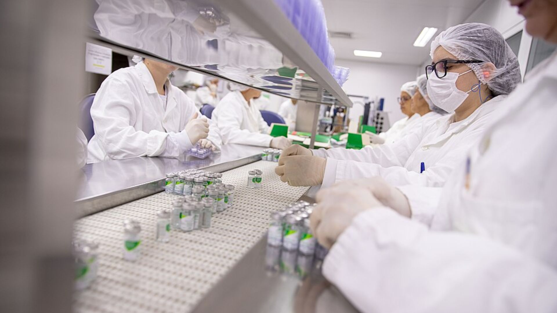 Lab workers in protective gear handle rows of small medicine vials on a pharmaceutical production line.