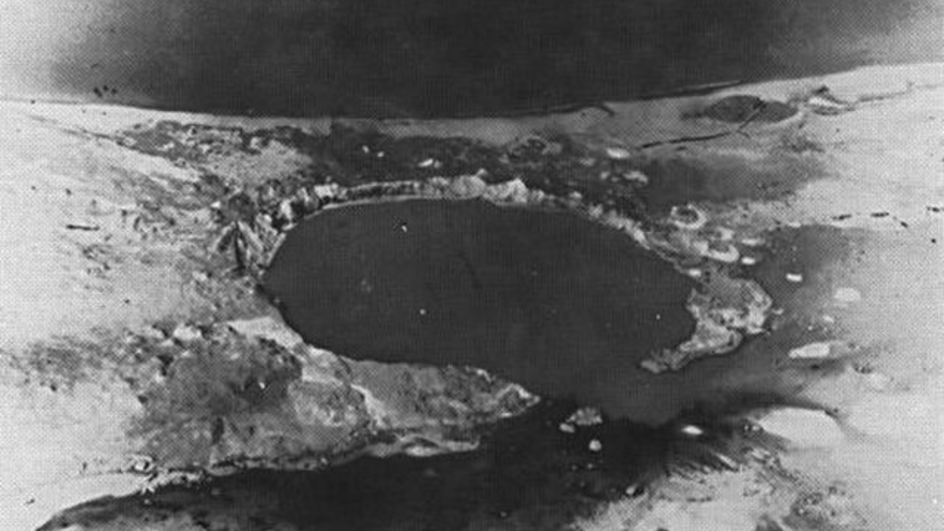 A historical black-and-white aerial photo showing a large crater formed by a nuclear explosion on an atoll.