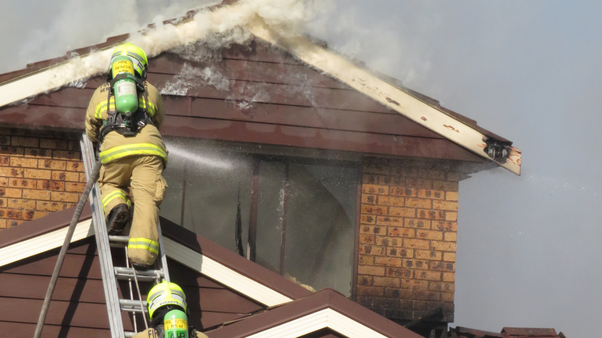 Two firefighters in full protective gear climb a ladder on the exterior of a brick house to extinguish a fire emitting thick white smoke from the roof.