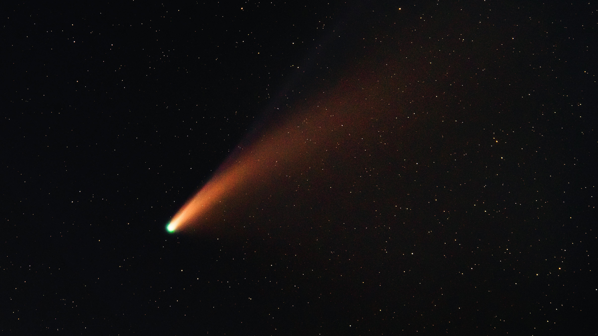 A comet with a bright green nucleus and a long, glowing orange dust tail streaks across a dark field of stars.