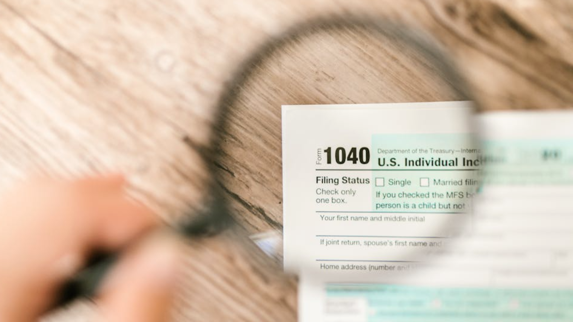 A hand holds a magnifying glass over a U.S. Individual Income Tax Return Form 1040, focusing on the _Filing Status_ section.