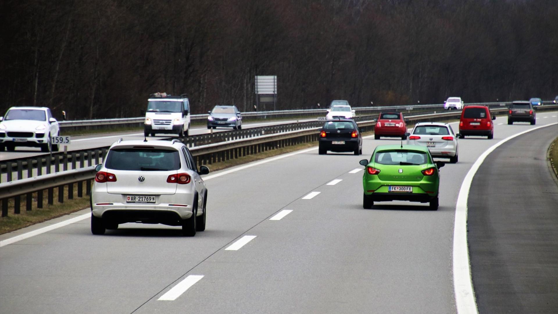 Several cars driving on a multi-lane highway bordered by guardrails and a forested area in the background under an overcast sky.