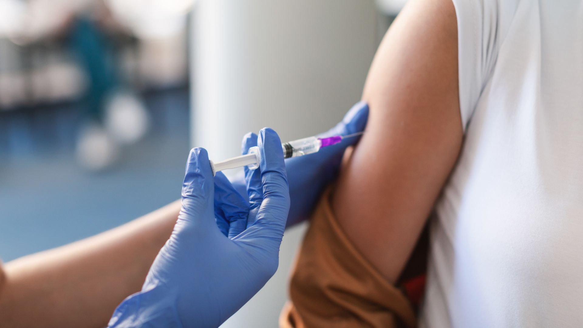 A gloved hand holds a syringe as a person receives a vaccine in the upper arm.