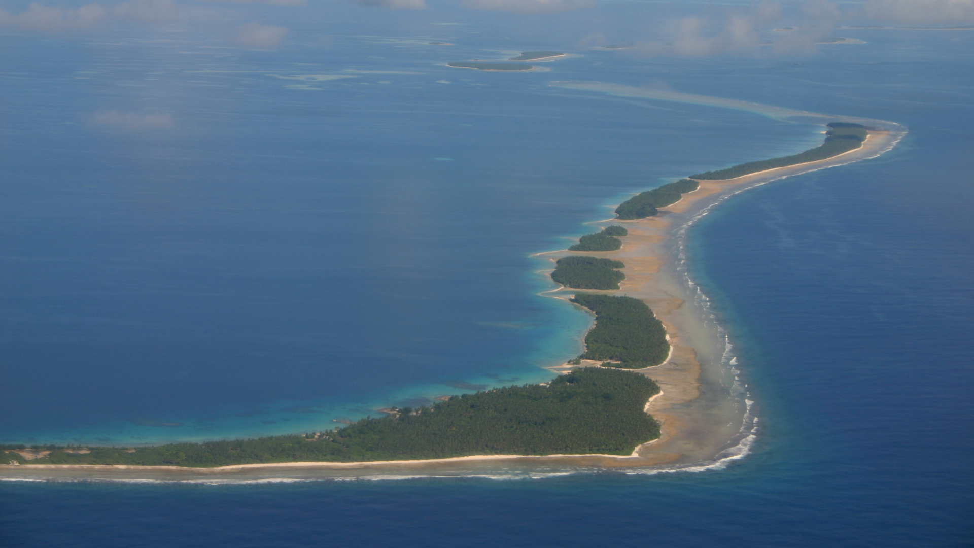 An aerial view of a long, narrow tropical atoll with green vegetation surrounded by deep blue ocean water.