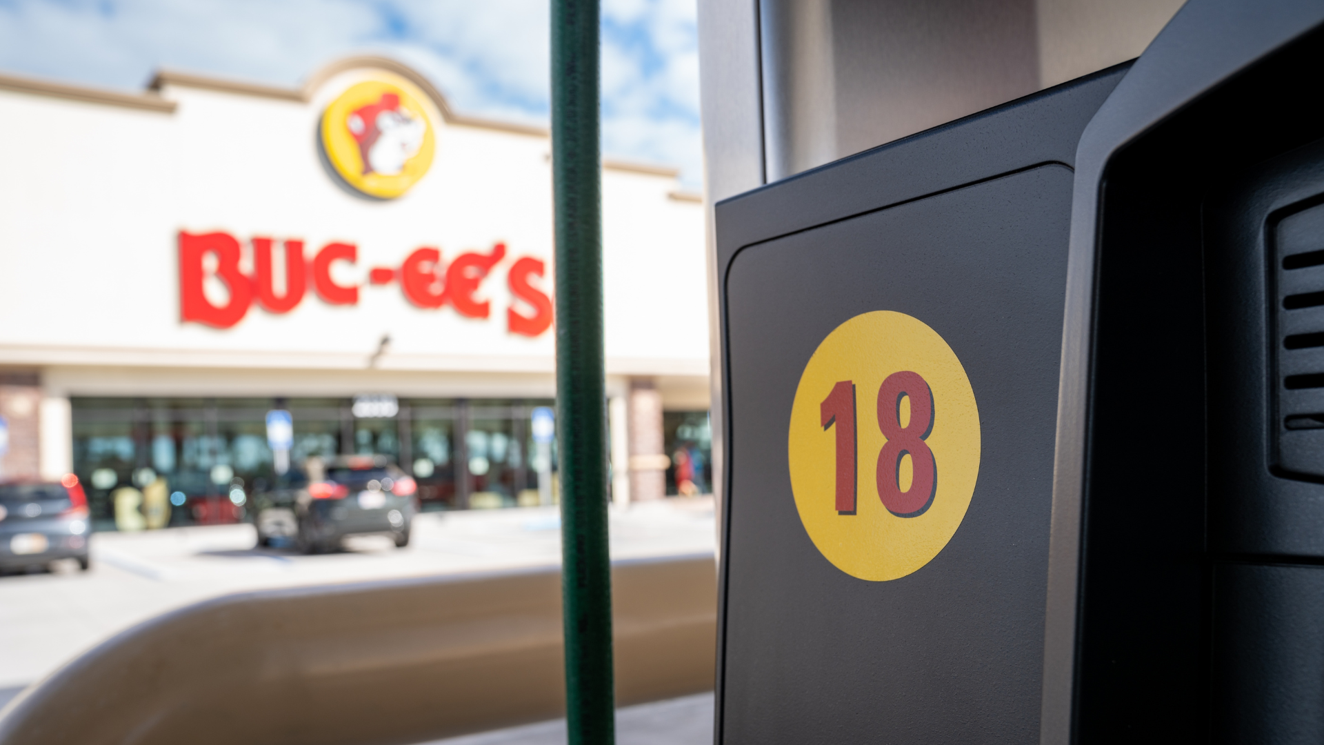 A close-up of a black gas pump with a yellow circle containing the number _18,_ with a blurred Buc-ee's convenience store and travel center in the background.