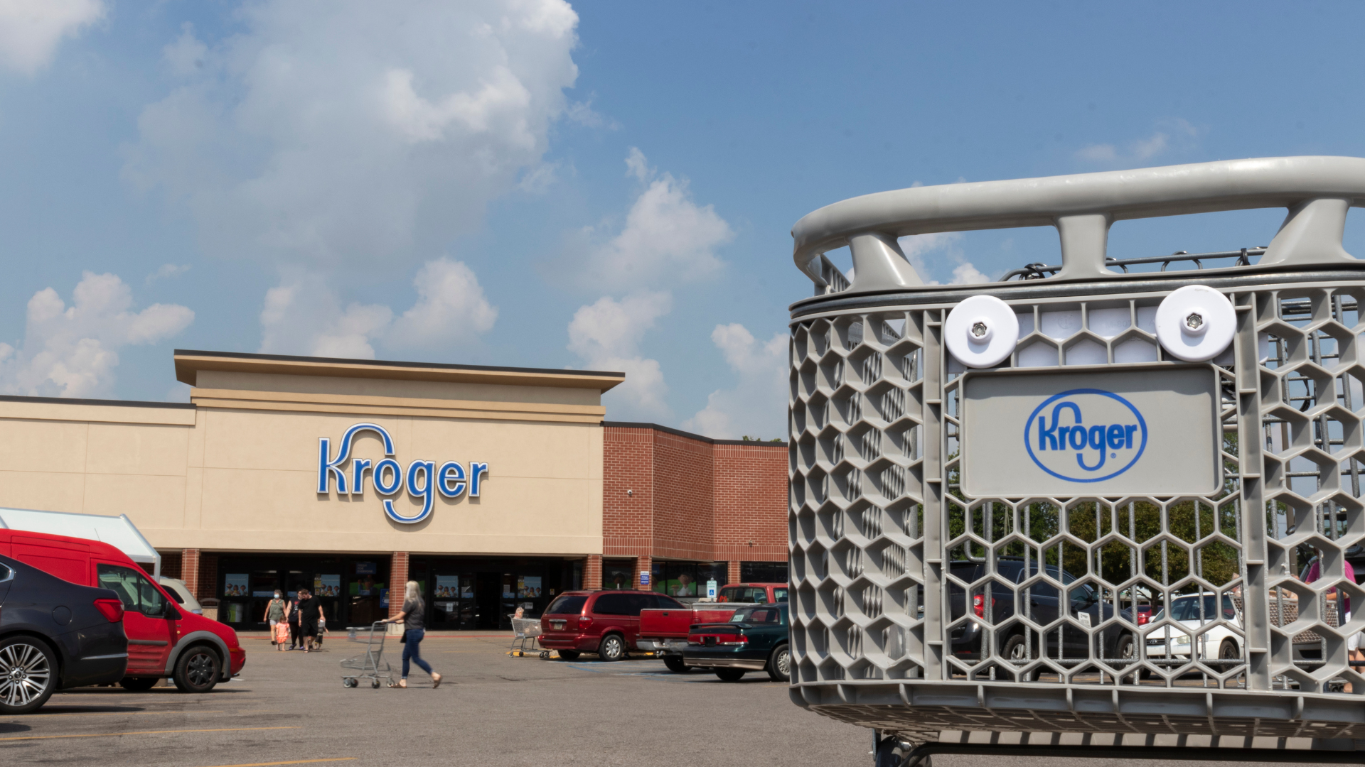 Kroger cart on the parking lot with the Kroger store in the background with people walking.